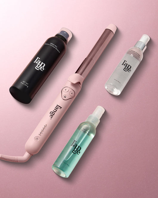 Pink curling wand alongside black, clear, and turquoise hair care bottles arranged on a soft pink background.