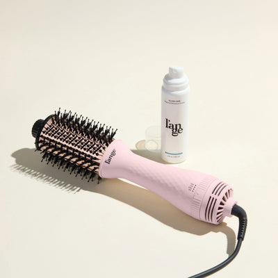 The Effortless Blowout Duo Value Set, featuring a Blush Le Volume brush and Glass Hair spray, displayed against a beige background