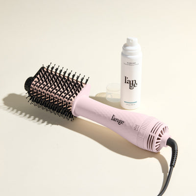 The Effortless Blowout Duo Value Set, featuring a Blush Le Volume Eleve brush and Glass Hair spray, displayed against a beige background
